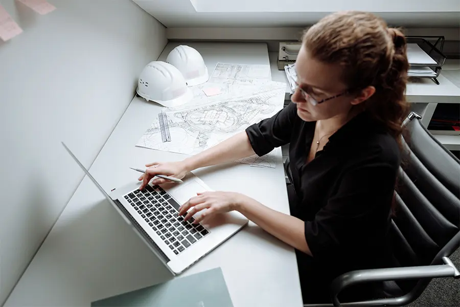 Construction IT support of a woman working on a laptop with construction plans and helmets laid out on her desk. Photo by Thirdman : https://www.pexels.com/photo/a-busy-woman-working-5582872/